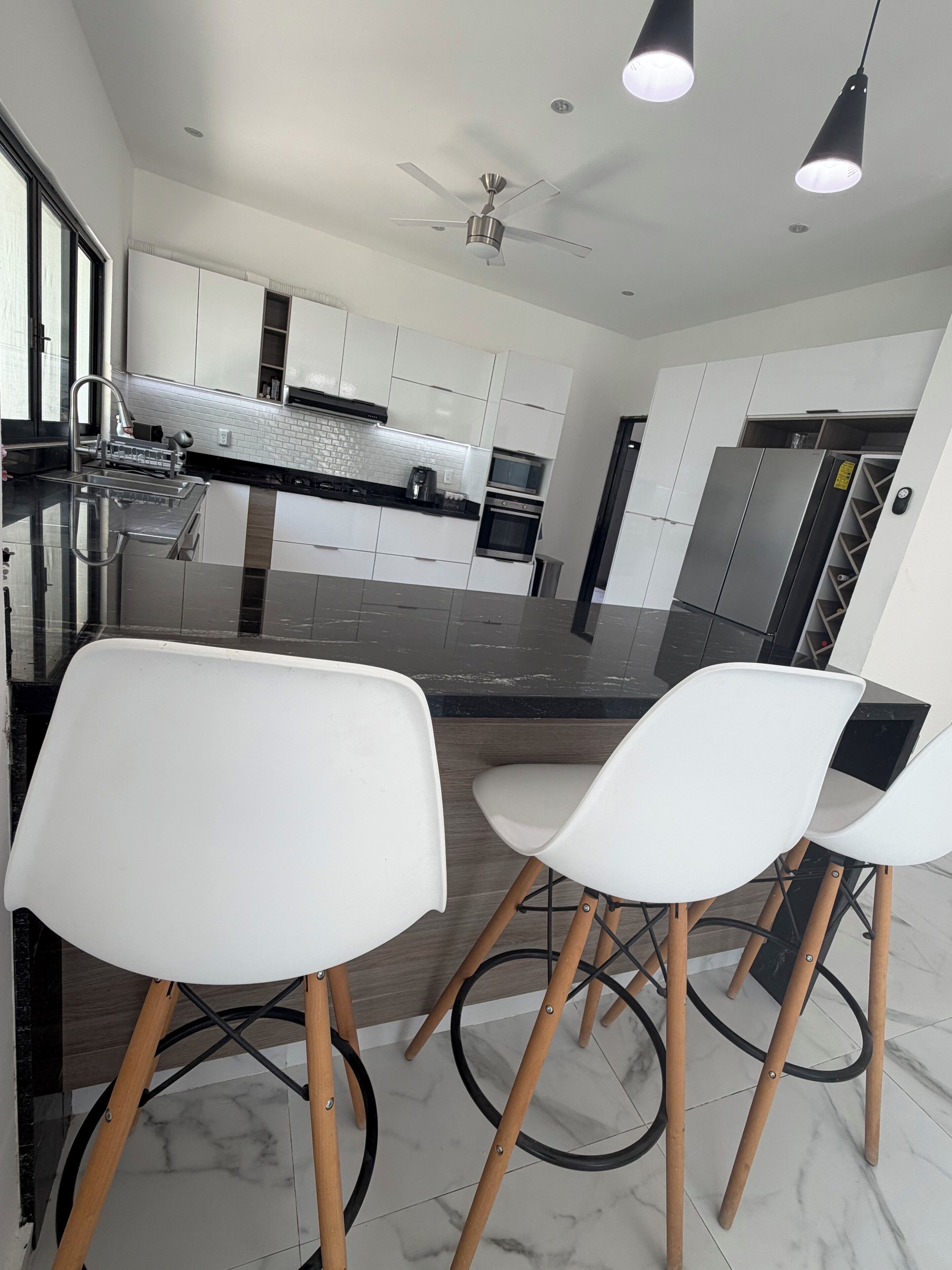 A modern kitchen with white cabinets, a stainless steel refrigerator, and a built-in oven. There are two white bar stools with wooden legs.