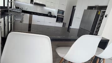 A modern kitchen with white cabinets, a stainless steel refrigerator, and a built-in oven. There are two white bar stools with wooden legs.