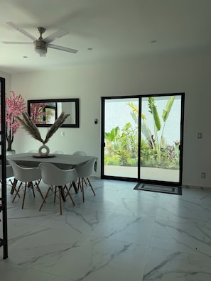 A dining area with a table, chairs, a ceiling fan, and a view of greenery through a glass door.