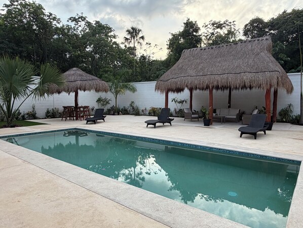 A pool area with lounge chairs and thatched umbrellas, surrounded by greenery.