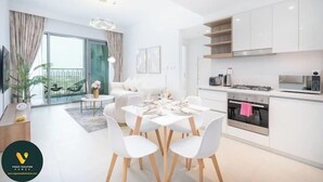 A modern kitchen with white cabinets, a dining area with a white table and chairs, and a large window with curtains. - Vogue | Downtown with Direct Access to Dubai Mall (Dubai)