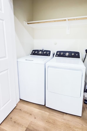A laundry room with two white front-loading washing machines and a wooden shelf above them. - Perfect 3-bedroom townhome located near Bush Airport (houston)