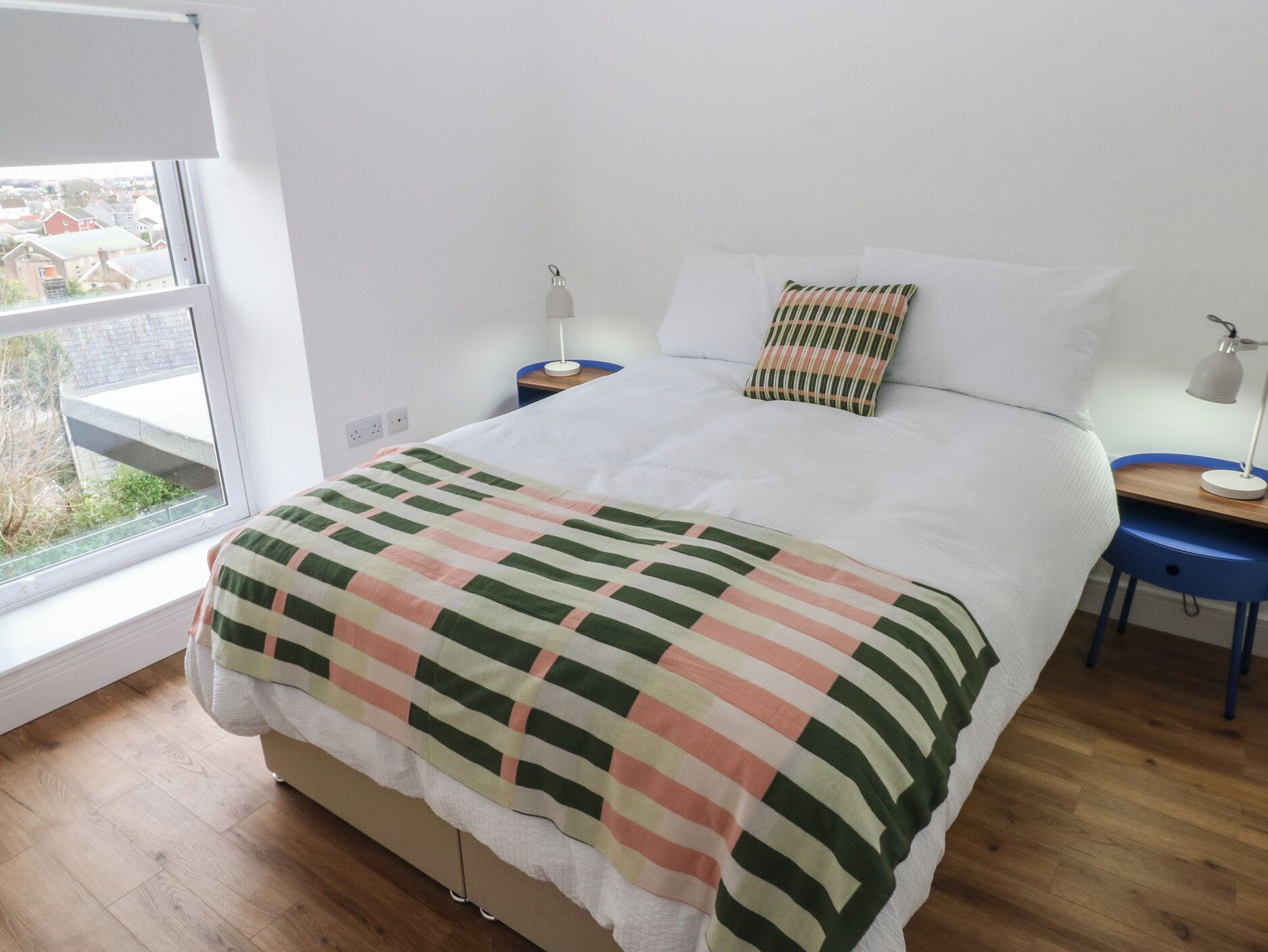 A bedroom with a bed, a window, a bedside table, a lamp, and a view of the outside.