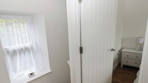 Ein Zimmer mit weißer Tür, einem Fenster mit durchscheinenden Vorhängen, einem Nachttisch mit Lampe und Holzboden.