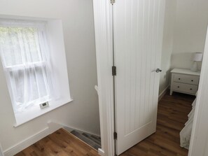 Ein Zimmer mit weißer Tür, einem Fenster mit durchscheinenden Vorhängen, einem Nachttisch mit Lampe und Holzboden.