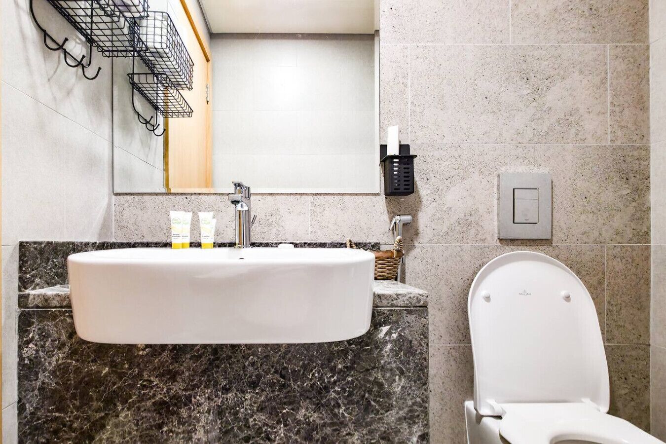 Ein Badezimmer mit einem weißen Waschbecken, einer Marmorarbeitsplatte und einer Toilette. Auf der Arbeitsplatte stehen Toilettenartikel und über dem Waschbecken ein Handtuchhalter.