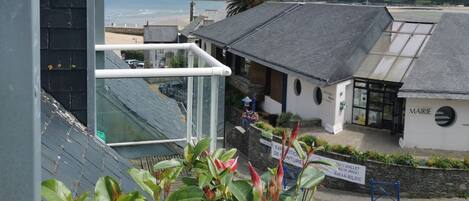 A coastal building with a balcony overlooking the sea, a sign indicating a summer exhibition, and a view of the beach and hills in the distance.