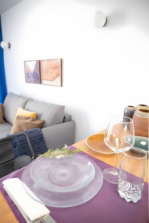 Ein Esstisch mit Glasplatte, ein Weinglas und ein Wasserglas, eine Couch mit Kissen sowie eine wandmontierte Lampe.
