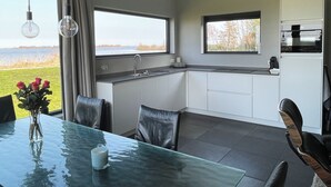 A modern kitchen with a view of the outdoors, a dining area with a glass table, and leather chairs. - Modern, spacious vacation apartment with a picturesque view of Lake Tjeukesee (Oosterzee)