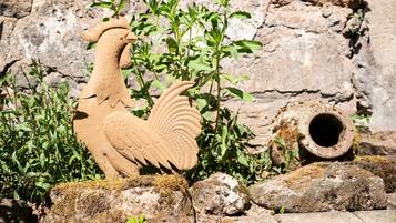 A rooster sculpture, a stone pipe, and greenery in a rustic outdoor setting.