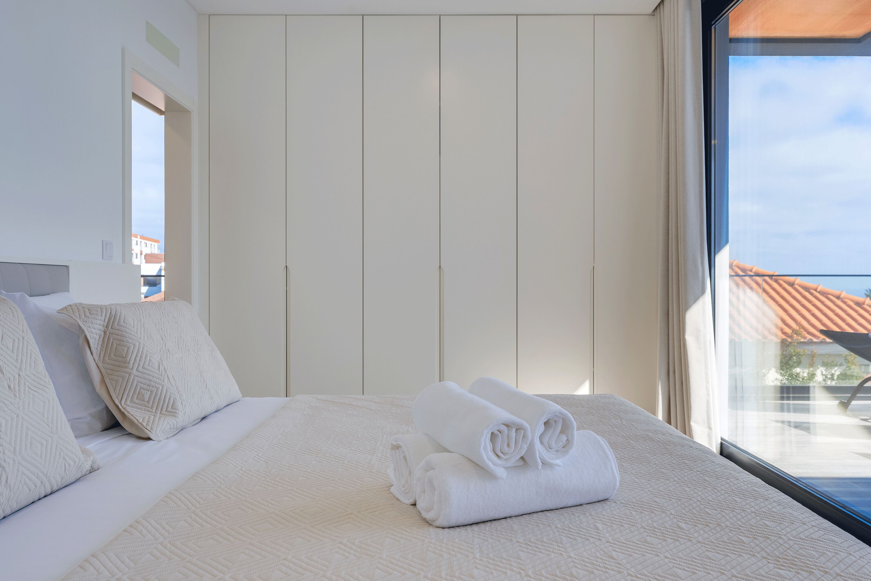 A neatly made bed with rolled white towels, a large window with a view of buildings, and a white wardrobe.