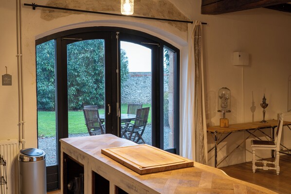 A kitchen with a wooden countertop, a table, and chairs. There is a window with a view of a garden and a stone wall. - House 3 on the Embourg estate (Souvigny)