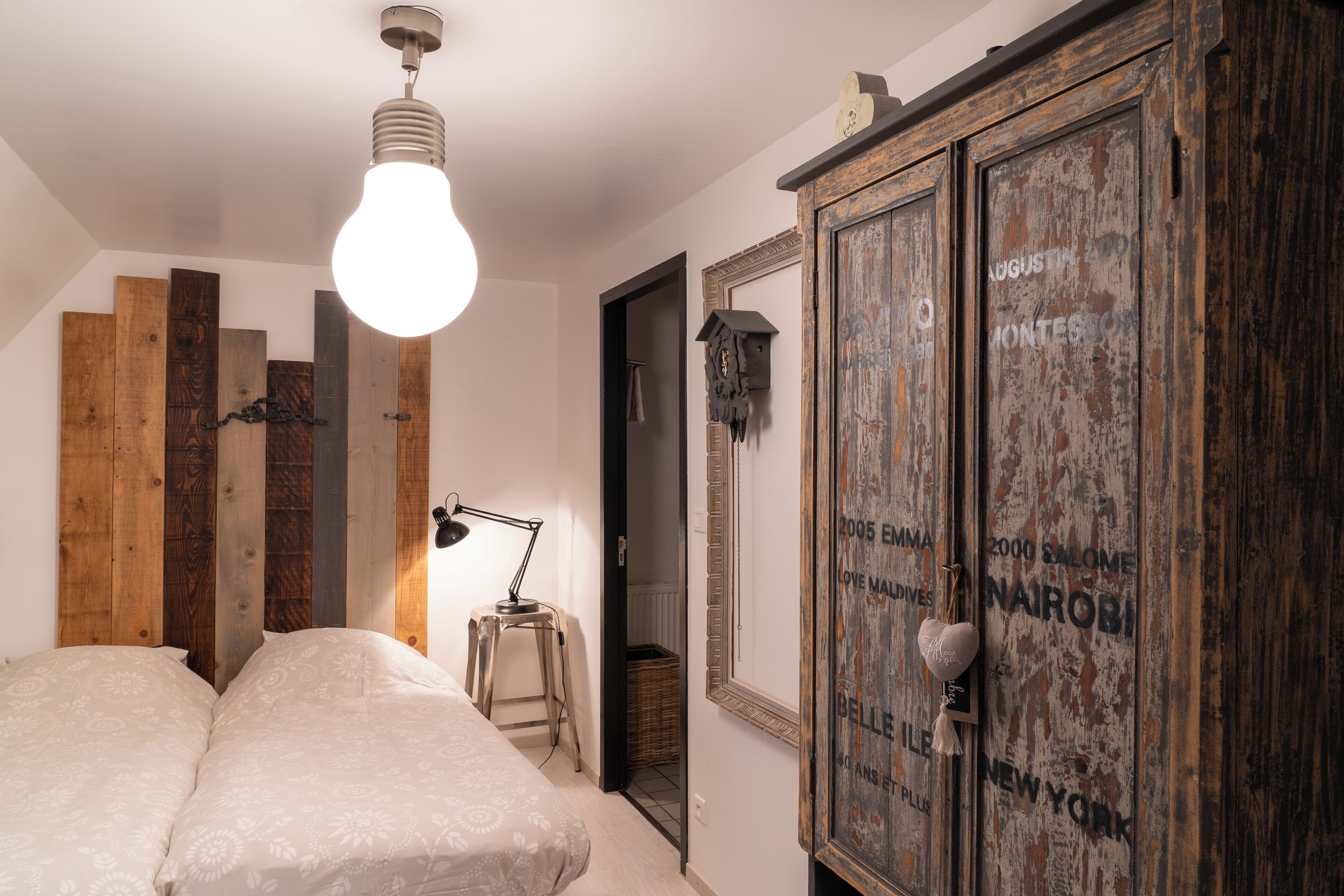 A bedroom with a bed, a wooden headboard, a hanging light fixture, and a wardrobe with travel destinations written on it.