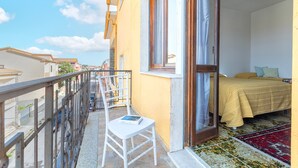 Ein Balkon mit Stuhl und Buch, ein Bett mit gelber Tagesdecke und ein gemustertes Teppich.