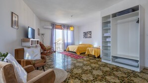 Ein Zimmer mit einer Couch, einem Sessel, einem Bett mit gelber Bettwäsche, einem Fernseher, einem Kleiderschrank und einem Fliesenboden.