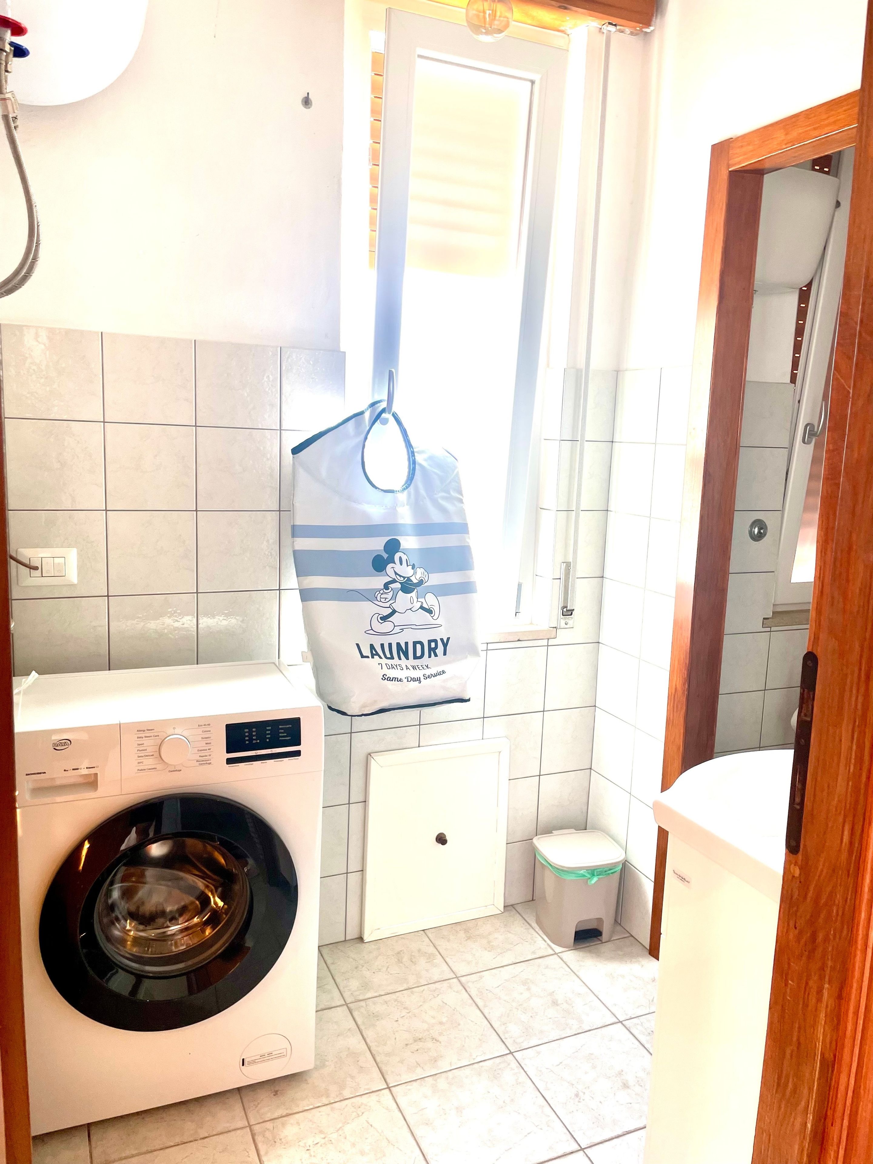 Une petite buanderie équipée d’un lave-linge à chargement frontal, d’un petit meuble blanc et d’un sac accroché au mur.
