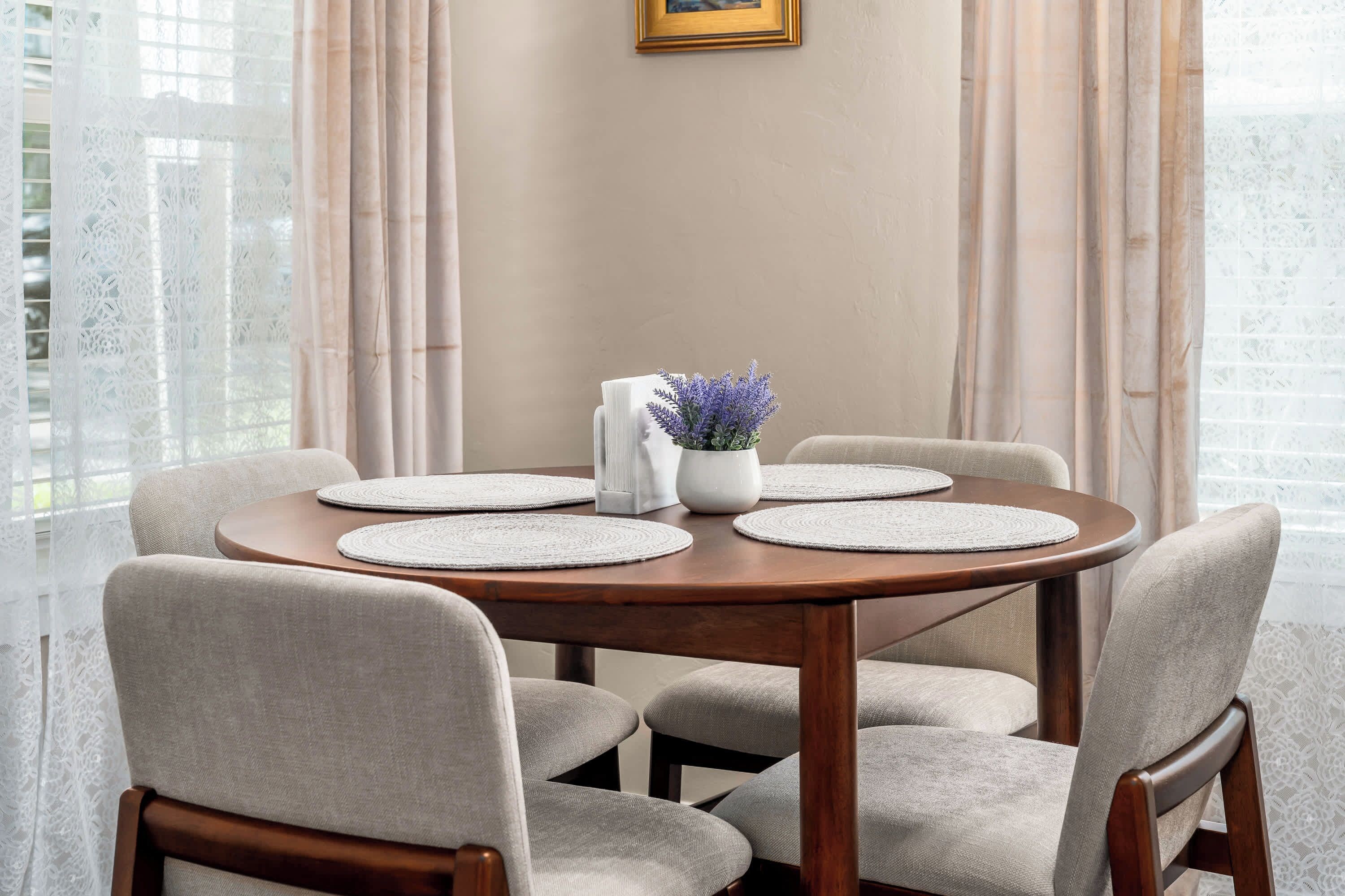 A round wooden table with four grey chairs, a white vase with purple flowers, and a framed picture on the wall.