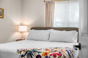 Un lit bien fait, avec du linge de lit blanc, un plaid à motifs floraux et une lampe de chevet.