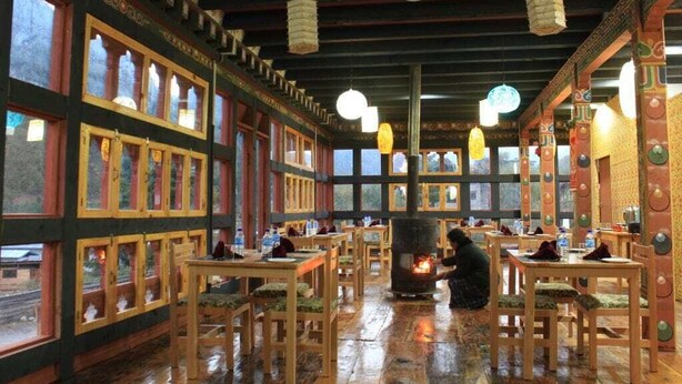 Un restaurant avec des tables et des chaises en bois, de grandes fenêtres et un poêle à bois central.