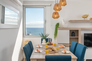 A dining area with a view of the sea, a table set with breakfast, and a TV on the wall. - Premium apartment panoramic seaview |AtlanticFrame (Câmara De Lobos)