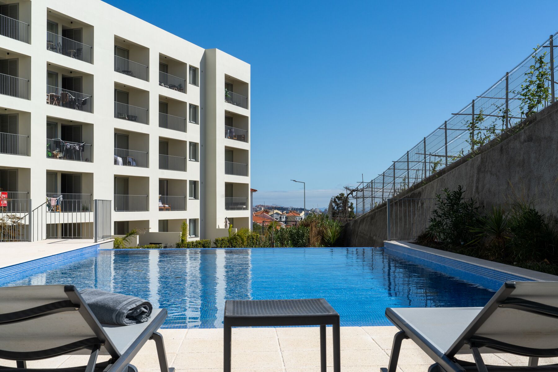 A modern building with multiple balconies, a swimming pool with lounge chairs, and a view of the sea.