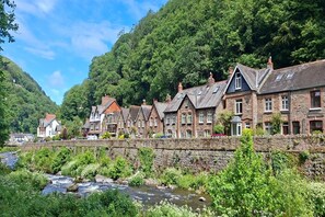 A row of stone buildings with a river in the foreground and a forested hill in the background. - A lovely 3 bed Riverside cottage in Lynmouth (Lynmouth)
