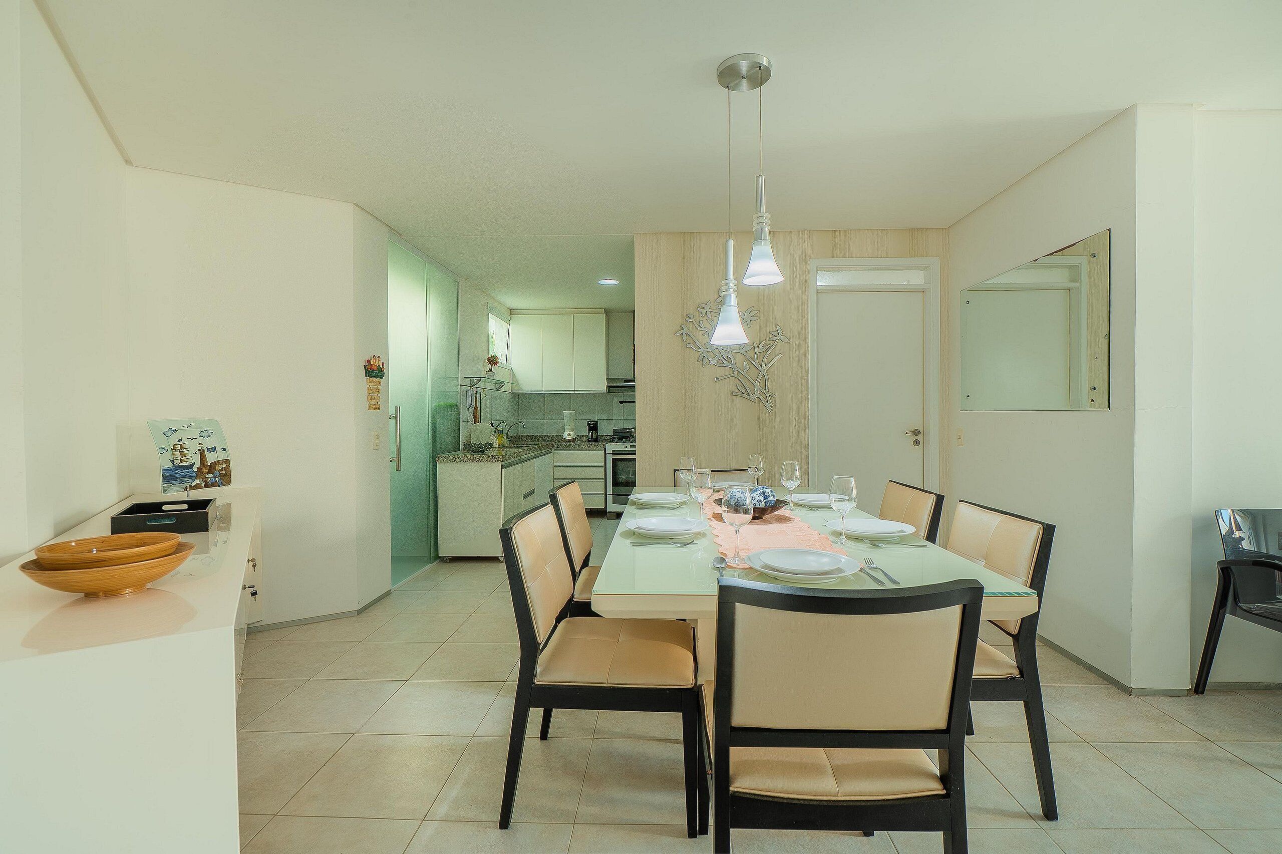 A dining area with a table set for four, chairs, a hanging light fixture, and a kitchen in the background.