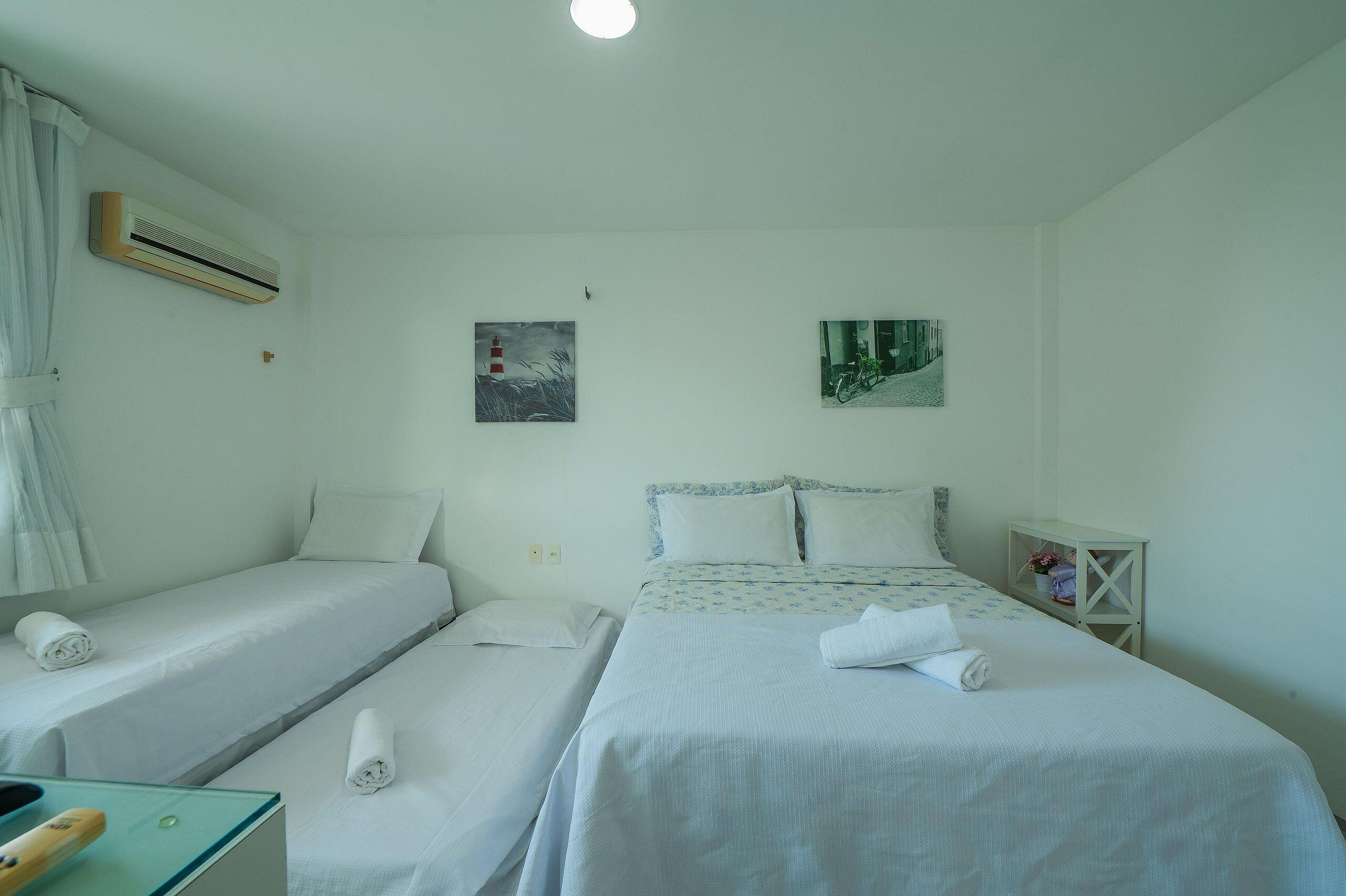 A hotel room with two single beds, a small table with a chair, an air conditioner, and two wall-mounted paintings.