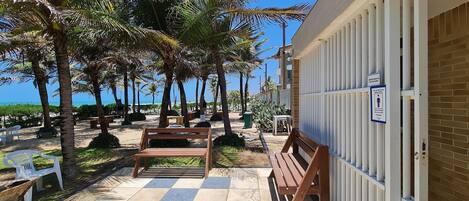 A checkered outdoor patio with wooden benches and a white plastic chair, palm trees, and a building with a white fence and a sign.