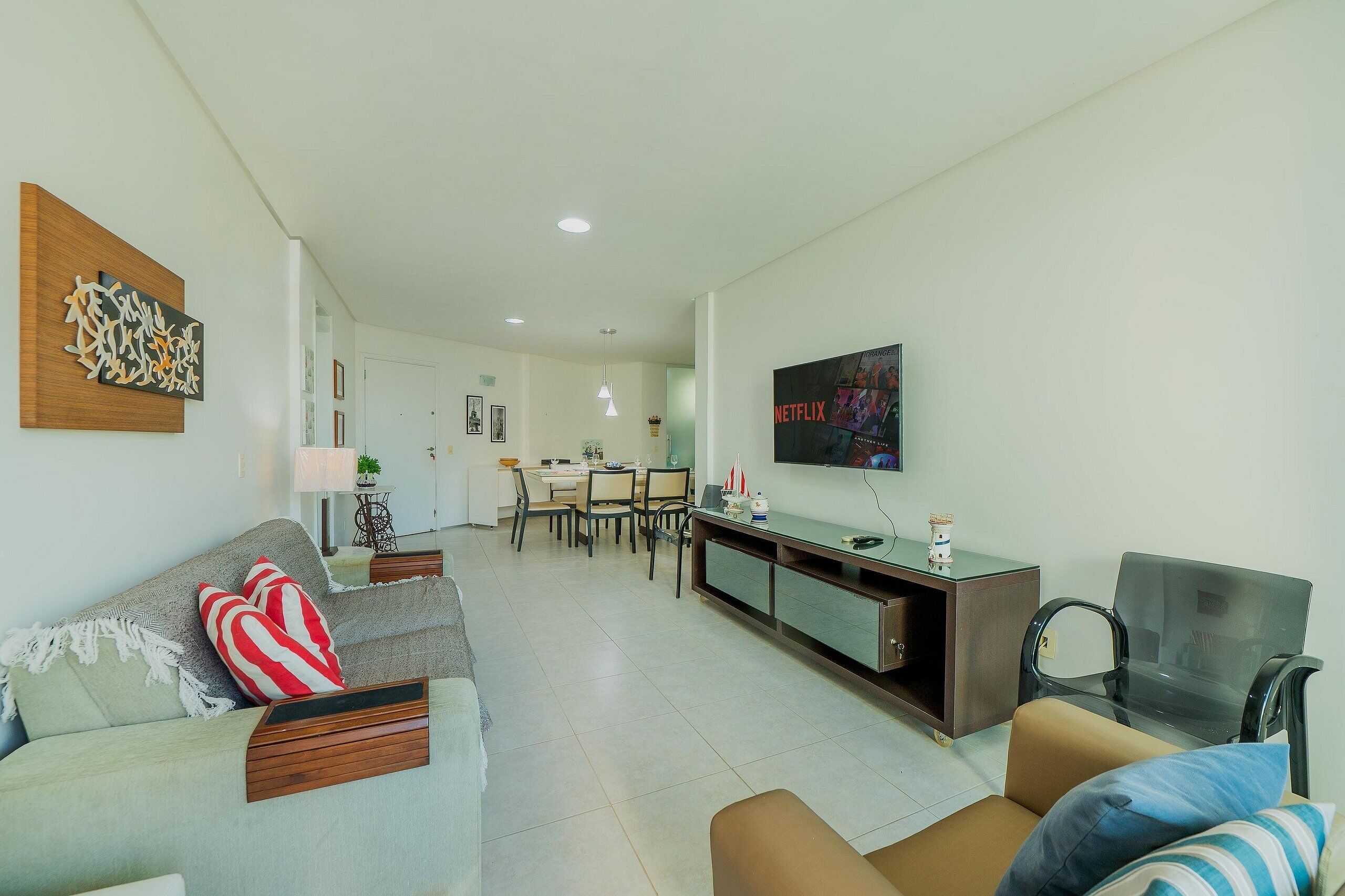 A modern living room with a sofa, armchairs, a dining area, and a TV mounted on the wall.