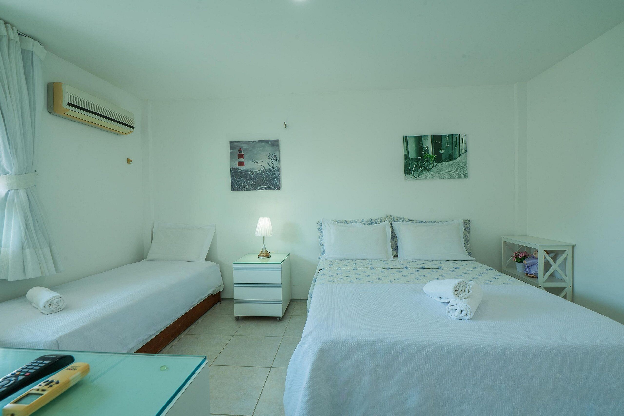A hotel room with two beds, a nightstand with a lamp, an air conditioner, and two wall-mounted pictures.