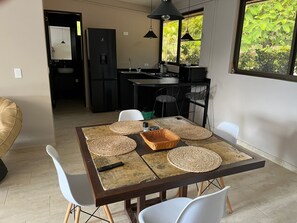 A dining area with a table set for four, surrounded by white chairs. A black refrigerator and a kitchen with a window are visible in the background.
