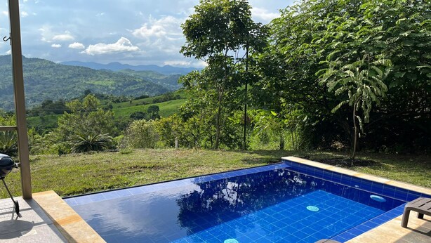 Ein rechteckiger Swimmingpool mit blauen Fliesen, umgeben von einer Terrasse mit Blick auf grüne Hügel und Bäume.