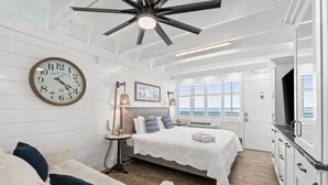 Ein Schlafzimmer mit einem Bett, Nachttischen, einer Uhr und Blick auf das Meer durch Fenster.