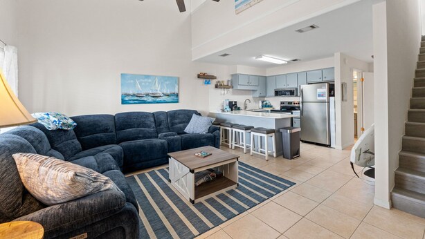 A modern living room with a dark blue sofa, a wooden coffee table, and a striped rug. A ceiling fan and a painting of a sailboat are visible. The kitchen area is equipped with stainless steel appliances and light blue cabinets. - Pet-Friendly 2BR | Walk to the Beach | Pool (Panama City Beach)