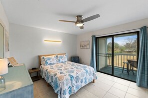A bedroom with a bed, a ceiling fan, a nightstand, a desk with a lamp, and a sliding glass door leading to an outdoor area. - Pet-Friendly 2BR | Walk to the Beach | Pool (Panama City Beach)