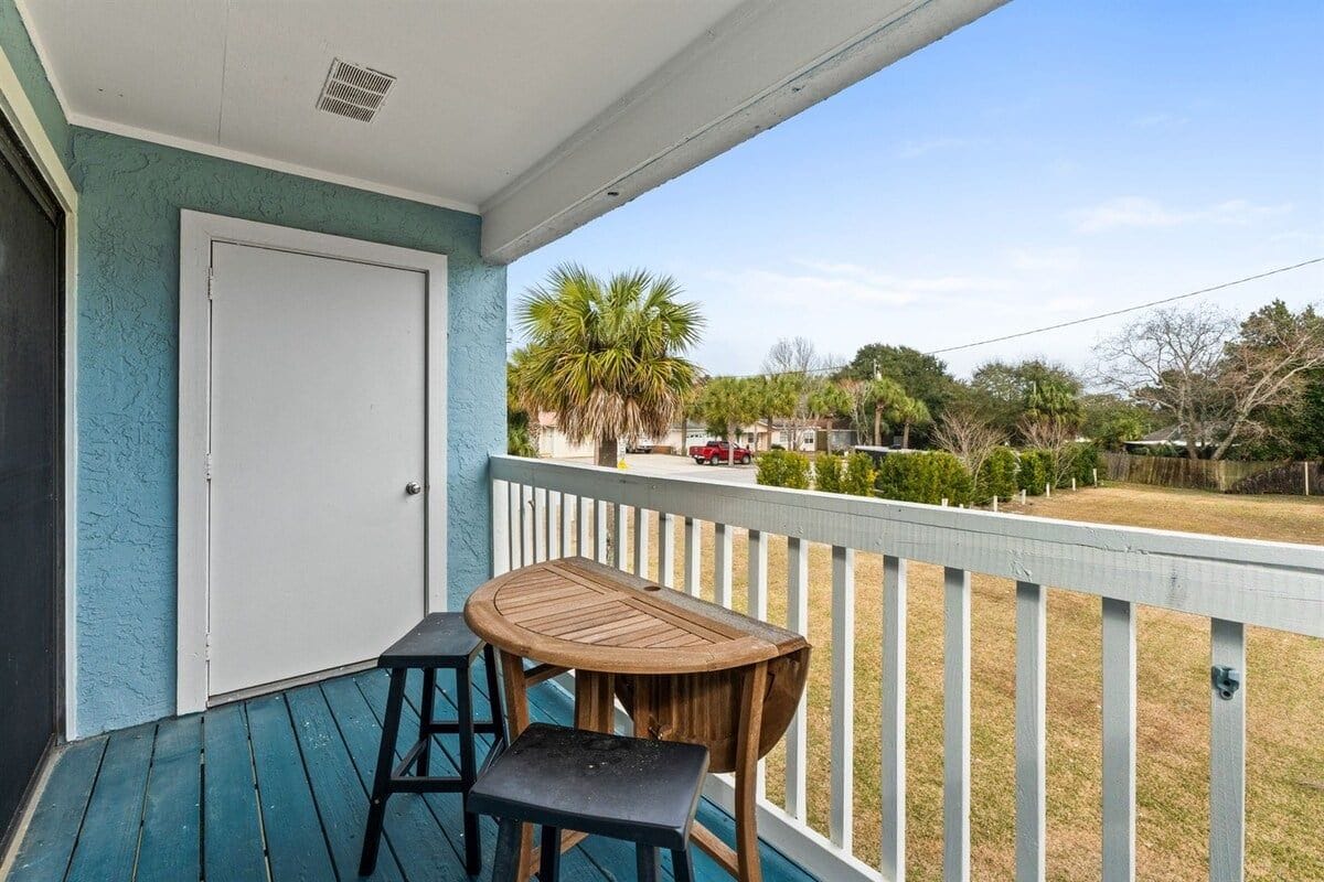 A covered porch with a table and chairs, a door, and a view of a residential area with trees and a parked car.