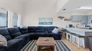 A modern living room with a dark blue sectional sofa, a wooden coffee table, and a striped rug. A painting of sailboats hangs on the wall. - Pet-Friendly 2BR | Walk to the Beach | Pool (Panama City Beach)