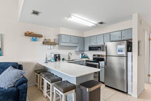 A compact kitchen with a central island, bar stools, a refrigerator, and a microwave. - Pet-Friendly 2BR | Walk to the Beach | Pool (Panama City Beach)
