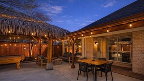 Une terrasse extérieure avec une table et des chaises en bois, une structure dotée d’un toit de chaume et des guirlandes lumineuses.
