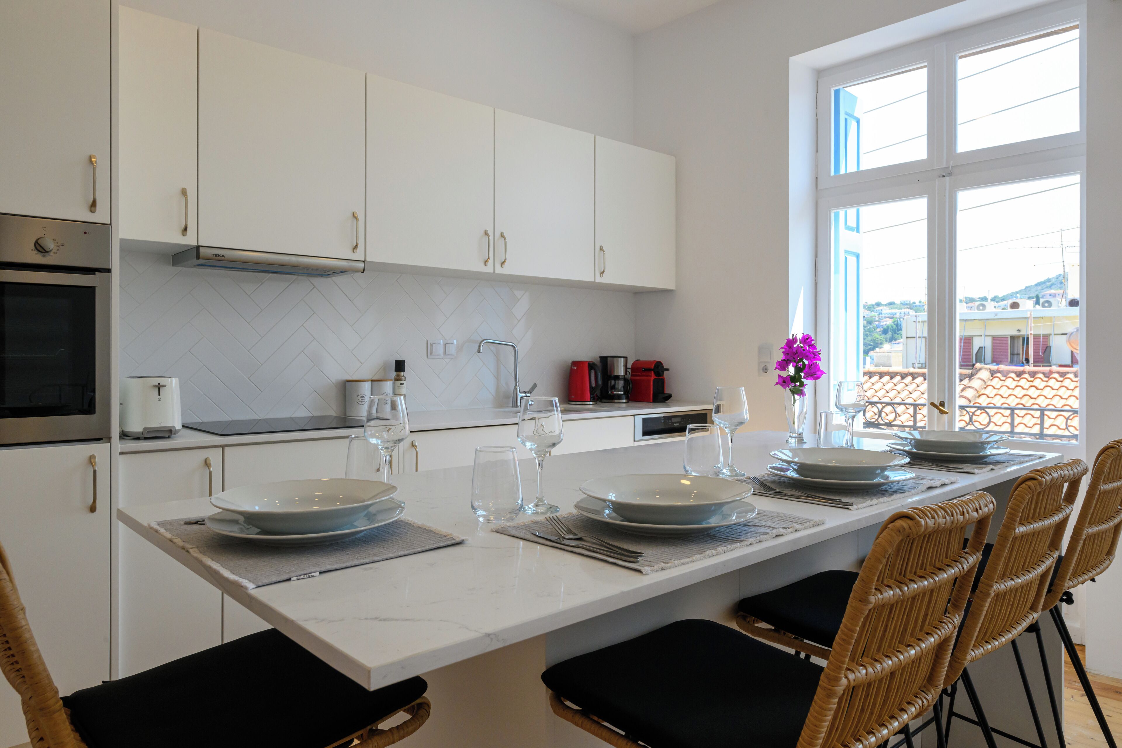 Une cuisine moderne avec des meubles blancs, un plan de travail en marbre et un coin repas comprenant des chaises en osier et une table blanche dressée pour quatre personnes.