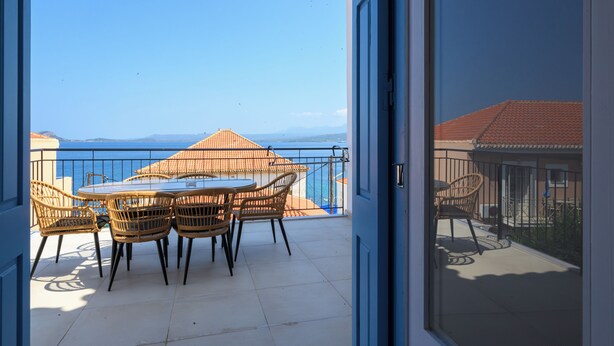A balcony with a table and chairs, a view of the sea, and a building with a red roof.