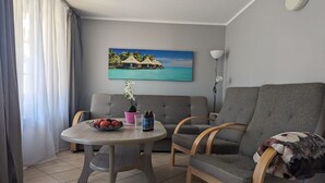 Una sala de estar con dos sofás grises, una mesa redonda de madera con un cuenco de fruta, un florero rosa y una botella. Un cuadro de una playa tropical adorna la pared.