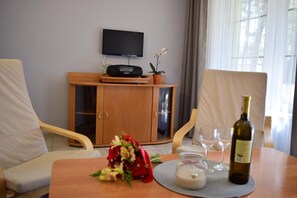 Hotellihuone, jossa on puinen kaappi, sen yläpuolella seinälle asennettu televisio, maljakko täynnä kukkia, pullo viiniä ja kaksi tuolia.