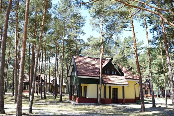Perinteinen puinen mökki, jossa on punatiilinen katto, ja sen ympärillä kohoaa korkeita mäntyjä.