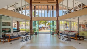 Een ruime lobby met een hoge plafond, grote ramen en uitzicht op het strand. Twee banken en een salontafel staan centraal.