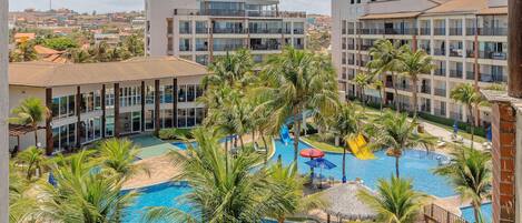 A resort with a central swimming pool surrounded by palm trees, multiple-story buildings, and a view of the surrounding landscape.
