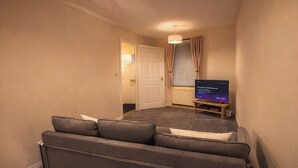 Ein kleiner, schwach beleuchteter Raum mit einem grauen Sofa, einem Fernseher auf einem Holzgestell und einer Tür, die in einen anderen Raum führt.
