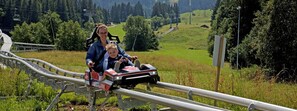 Eine Person und ein Kind auf einer Holzachterbahn in einer Berglandschaft.