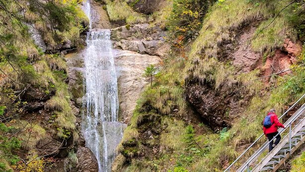 Uma pessoa caminhando sobre uma ponte de metal acima de uma cascata, em meio à floresta.